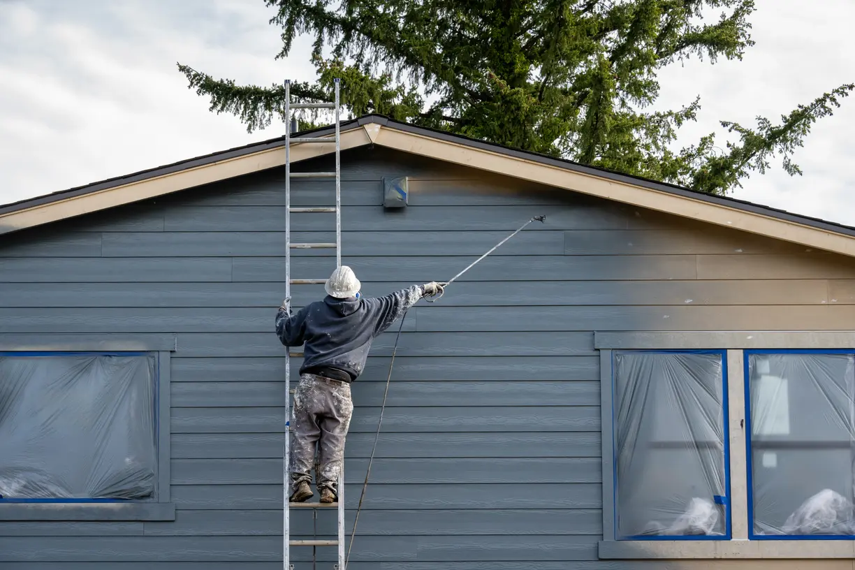 A painter on a ladder using a paint sprayer to paint the outside of a clients home